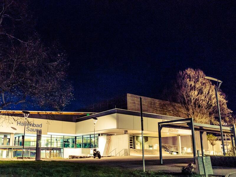 Das Bild zeigt ein beleuchtetes Hallenbad mit Sauna vor einem dunklen Nachthimmel, umgeben von Bäumen.
