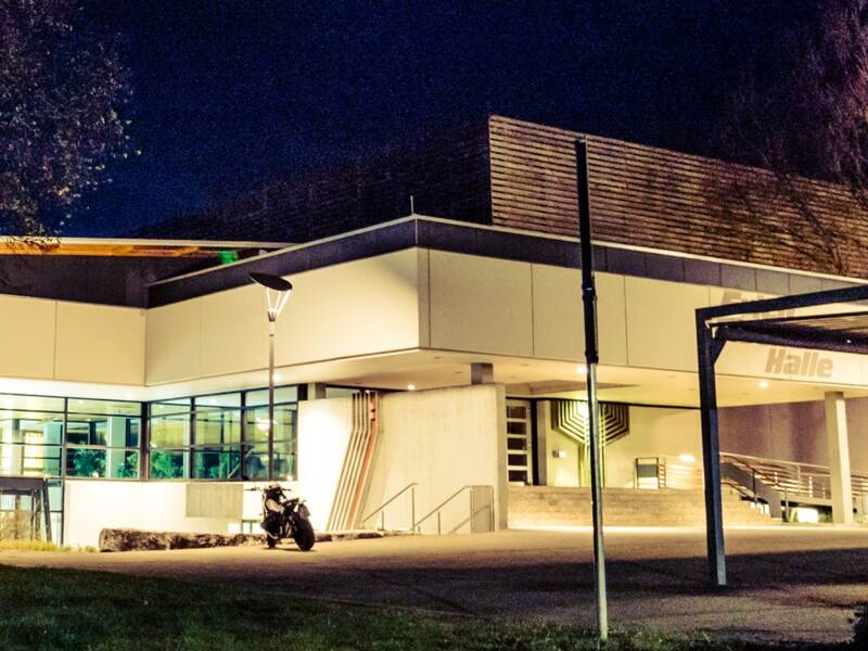 Das Bild zeigt ein beleuchtetes Hallenbad mit Sauna vor einem dunklen Nachthimmel, umgeben von Bäumen.
