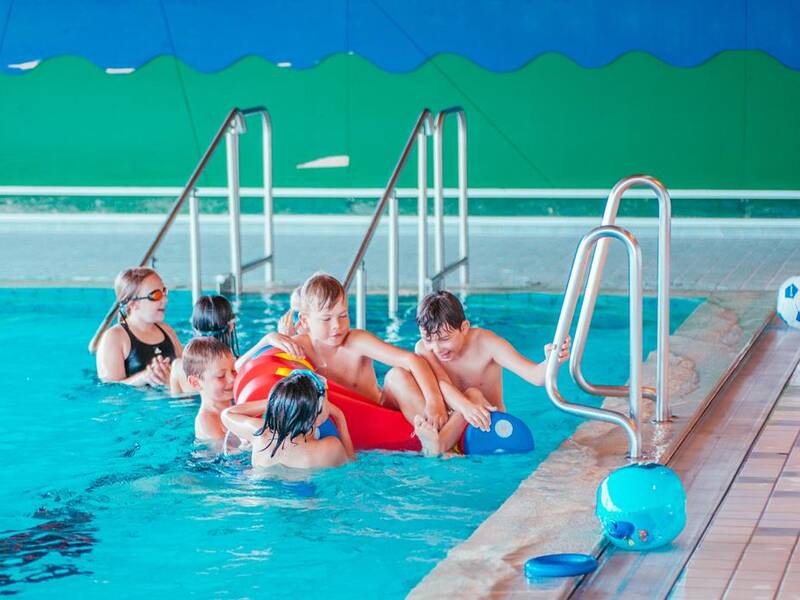 Mehrere Kinder spielen fröhlich im Wasser eines Hallenbads, umgeben von Badetieren und Schwimmhilfen.