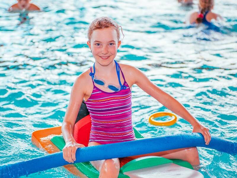 Ein lachendes Kind im gestreiften Badeanzug hält eine blaue Poolnudel, während es im Schwimmbecken sitzt.