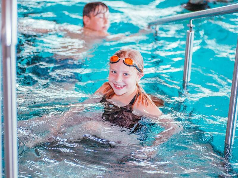 Ein Kind mit Schwimmbrille lächelt in einem Schwimmbecken, während eine weitere Person im Hintergrund schwimmt.