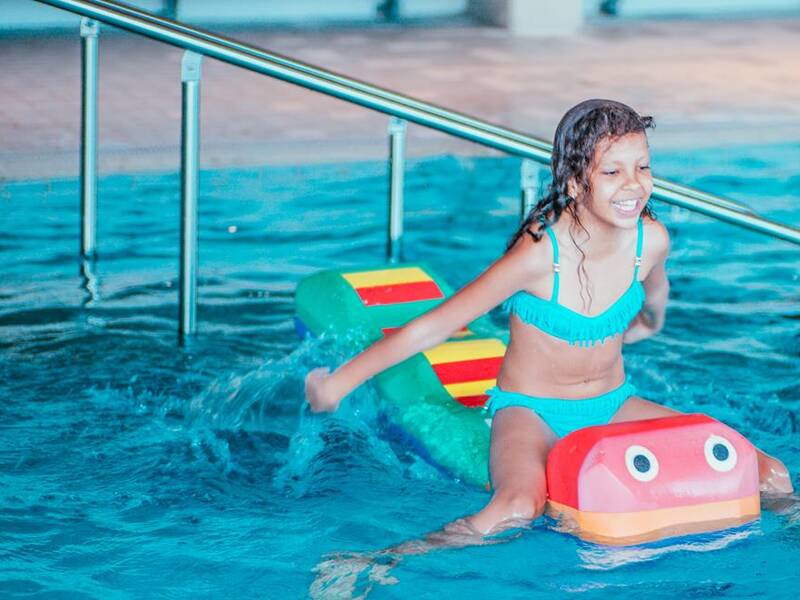 Ein Mädchen in einem türkisen Badeanzug sitzt lächelnd auf einem bunten Wasserspielzeug im Pool.