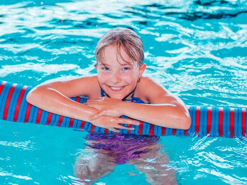 Ein Kind lächelt fröhlich, während es sich auf einer bunten Schwimmnudel im Wasser abstützt.