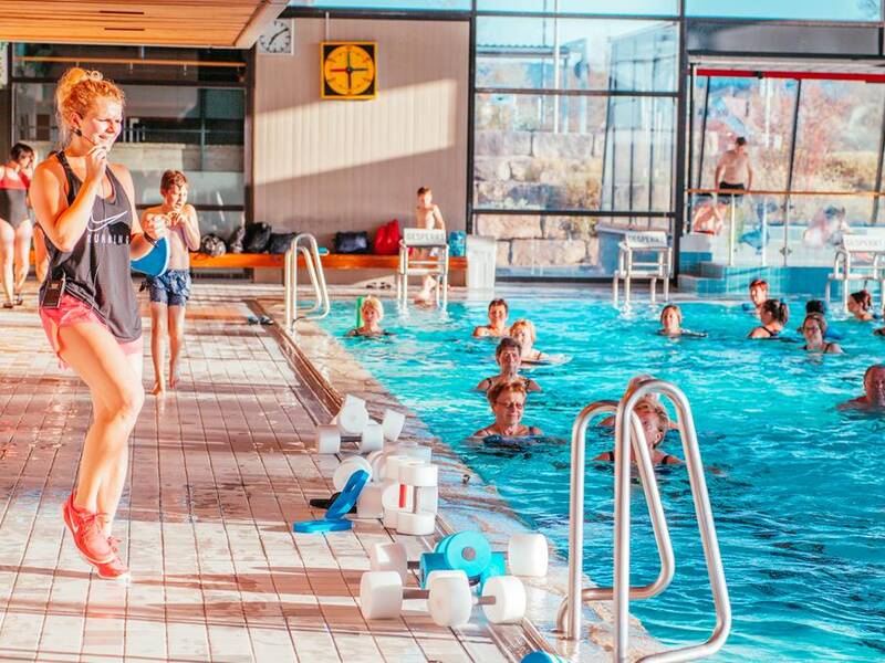 Eine Frau leitet eine Wassergymnastikstunde in einem Hallenbad, während mehrere Personen im Wasser mitmachen.