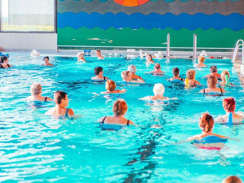 Eine Gruppe älterer Menschen nimmt an einer Aquafitness-Stunde in einem hellen Hallenbad teil.