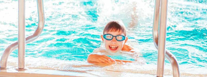 Ein fröhliches Kind mit Schwimmbrille genießt das Wasser in einem Pool, während es sich an der Kante festhält.