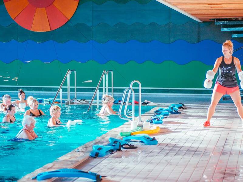 Eine Gruppe älterer Menschen macht Wassergymnastik in einem Schwimmbecken, während eine Trainerin am Beckenrand Übungen mit Hanteln vorführt.