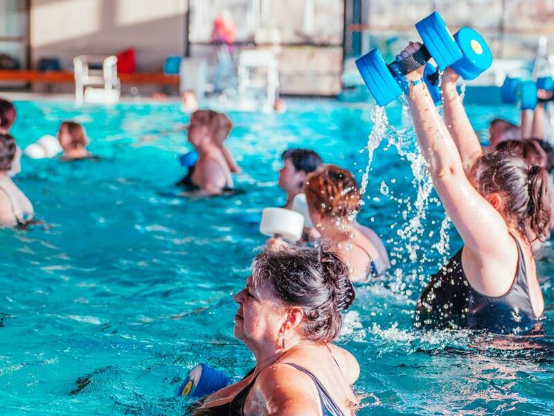 Mehrere Personen führen Wassergymnastik mit blauen Schaumstoffhanteln in einem Schwimmbad durch.