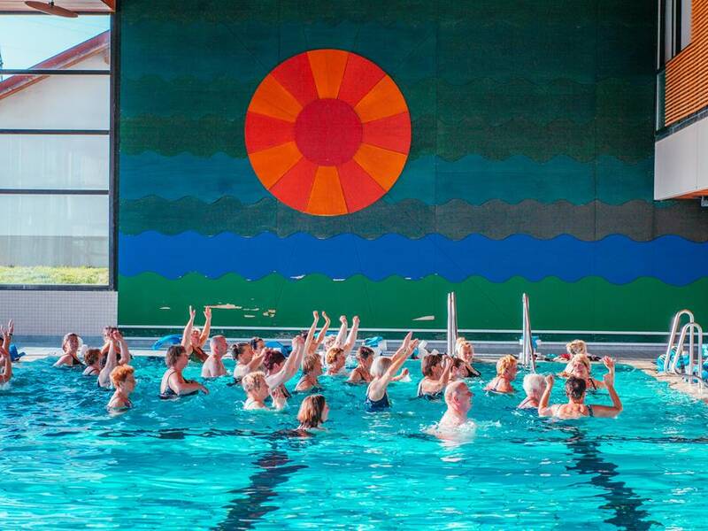 Eine Gruppe von Menschen macht Wassergymnastik in einem Hallenbad vor einer bunt gestalteten Wand mit einem großen, orangen Kreis.