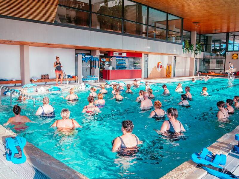 In einem Hallenbad macht eine Gruppe von Menschen gemeinsam Wassergymnastik unter Anleitung einer Trainerin am Beckenrand.