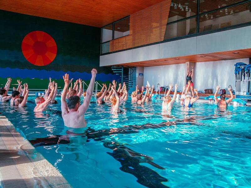 Eine Gruppe von Menschen macht Wasserübungen in einem Hallenbad, während ein Trainer sie anleitet.