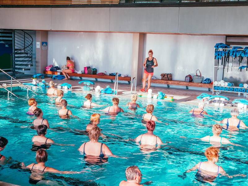 Eine Gruppe von Menschen nimmt an einem Wasser-Aerobic-Kurs in einem Hallenbad teil, während eine Trainerin am Beckenrand Anweisungen gibt.