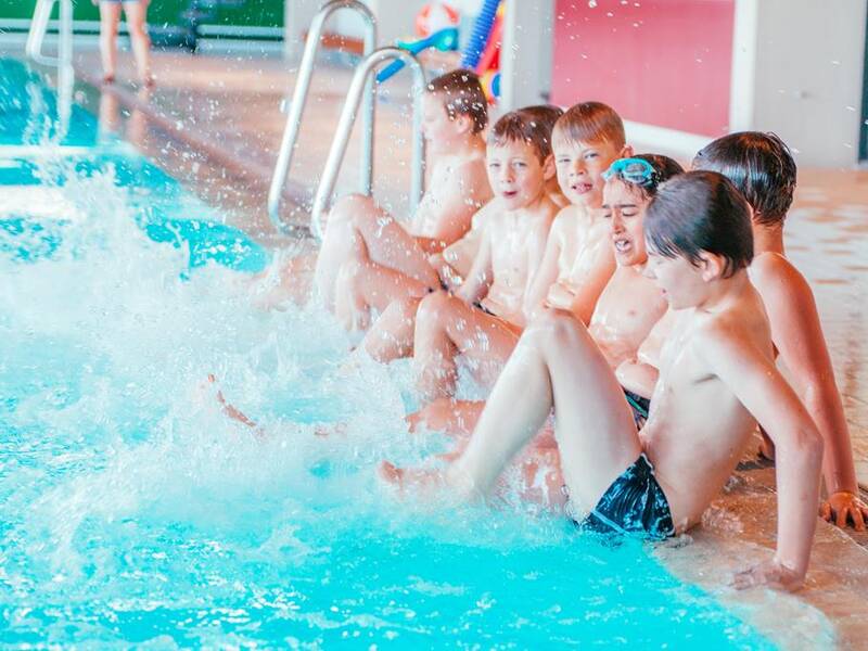 Eine Gruppe von Kindern sitzt am Beckenrand eines Schwimmbads und platscht mit den Beinen im Wasser.