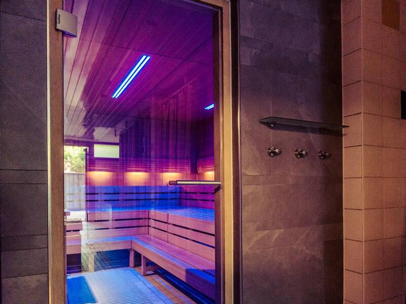 Eine Sauna mit Holzbänken und bunten LED-Leuchten ist durch eine Glastür sichtbar.