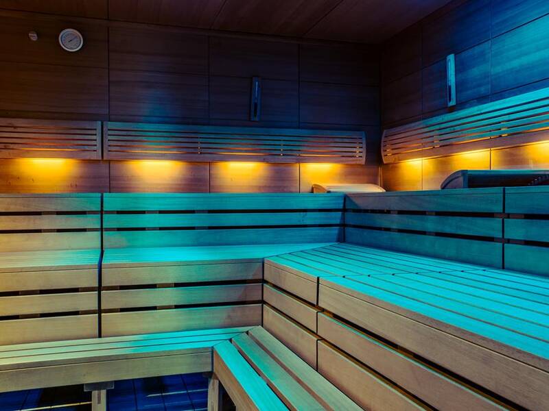 Die Sauna zeigt hölzerne Bänke in mehreren Ebenen, die von warmem gelbem und blauem Licht beleuchtet werden.